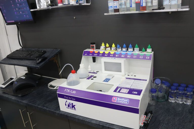 Una mesa con una pantalla y teclado de ordenador pegada a la pared, y a la derecha una maquina de análisis de agua de piscina